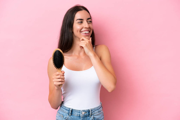 Young caucasian woman with hair comb isolated on pink background thinking an idea and looking side - Φωτογραφία, εικόνα