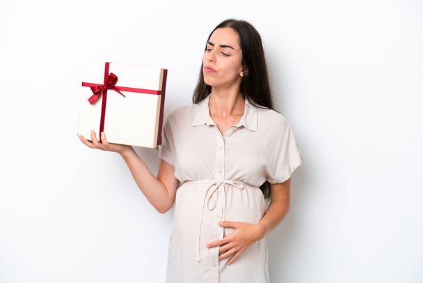 Beyaz arka planda izole edilmiş genç beyaz bir kadın hamile ve elinde şüpheleri varken bir hediye tutuyor. - Fotoğraf, Görsel