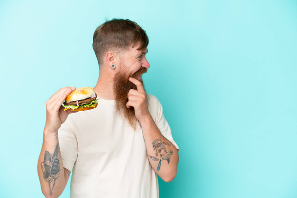 Рудий чоловік з довгою бородою тримає бургер ізольовано на синьому фоні, думаючи про ідею і виглядаючи збоку
 - Фото, зображення
