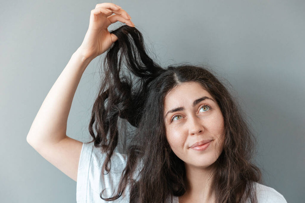 Νεαρή αναστατωμένη γυναίκα κρατά μια μπερδεμένη τούφα από τα μαλλιά της σε αμηχανία. Η έννοια της τριχολογίας και σμηγματορροϊκής δερματίτιδας - Φωτογραφία, εικόνα