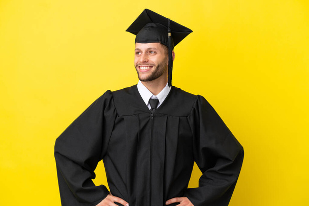 Young university graduate caucasian man isolated on yellow background posing with arms at hip and smiling - Fotografie, Obrázek