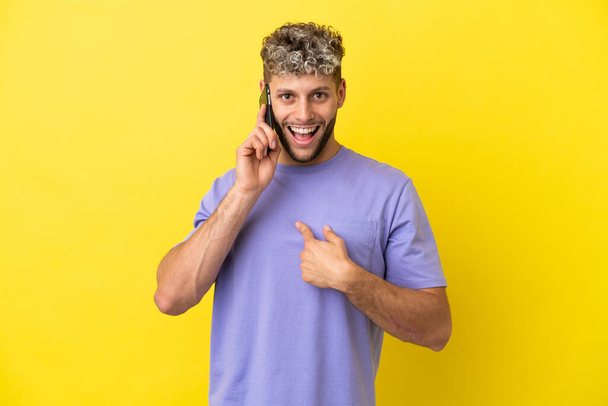 Молодой кавказский мужчина с мобильным телефоном на желтом фоне с неожиданным выражением лица - Фото, изображение