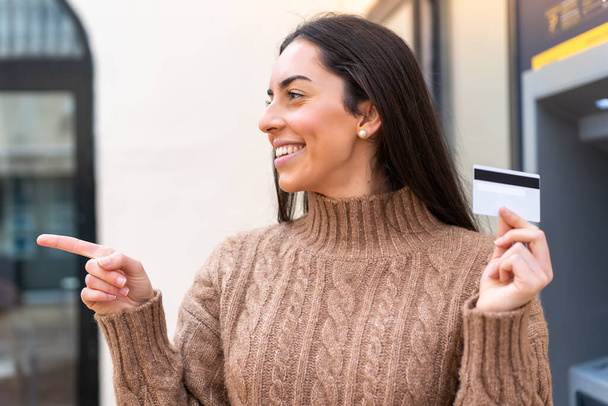 Mujer joven sosteniendo una tarjeta de crédito al aire libre apuntando hacia un lado para presentar un producto - Foto, Imagen