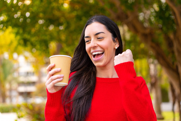 Młoda kobieta trzyma kawę na świeżym powietrzu, świętując zwycięstwo. - Zdjęcie, obraz