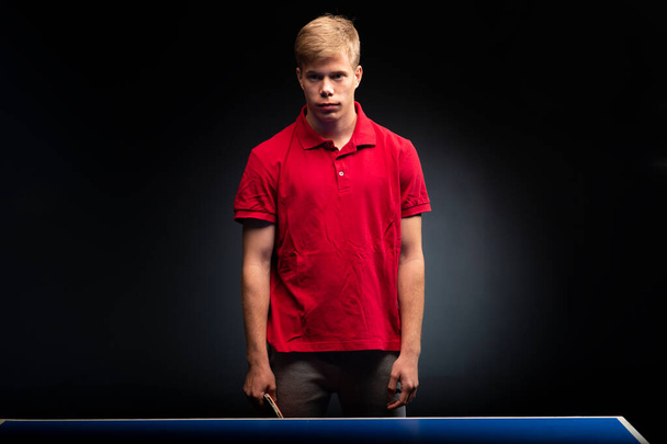 Portrait of a young man standing next to a table tennis table with a racket in his hand on a black background  - Zdjęcie, obraz