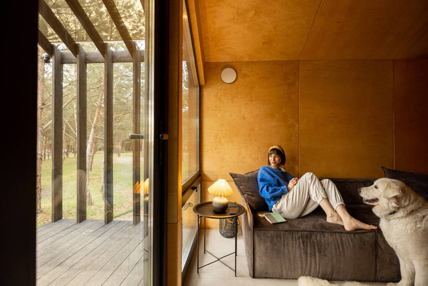 Woman reads a book while sitting relaxed on a couch by the window with her dog at wooden cottage in forest. Concept of home comfort and rest in a house on nature. Wide angle view - Fotoğraf, Görsel