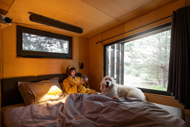 Kobieta używa telefonu leżąc w łóżku ze swoim słodkim psem w małej sypialni drewnianej kabiny na łonie natury - Zdjęcie, obraz