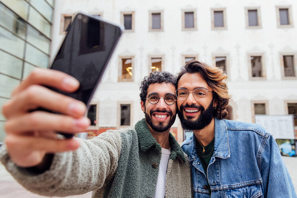 happy couple of gay men laughing taking a selfie photo with a mobile phone in the street, concept of urban lifestyle and love between people of the same sex - Foto, imagen