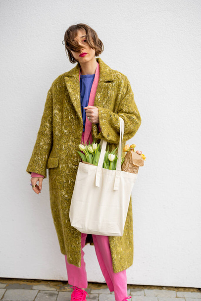 Portret stylowej kobiety w jasnych ubraniach z eko torebką pełną kwiatów na białym tle. Torba płócienna z czystą przestrzenią - Zdjęcie, obraz