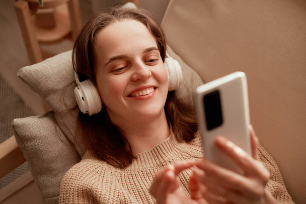 Photo of young adult Caucasian woman wearing headphones holding cell phone while lying on sofa in her apartment, using mobile phone for checking social networks or chatting in hands. - Fotografie, Obrázek