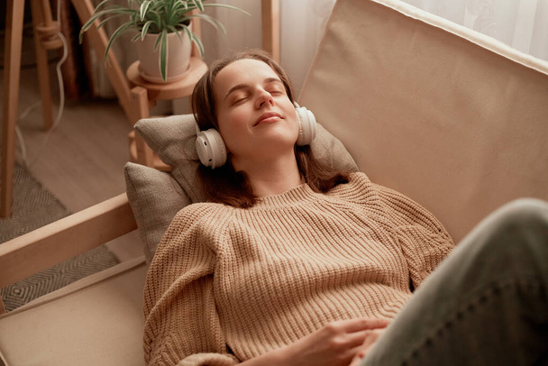 Happy Caucasian woman lying on sofa and listening to music in headphones, enjoying audio playing in earphones, relaxing and chilling at home, expressing positive emotions having fun while laying along - 写真・画像