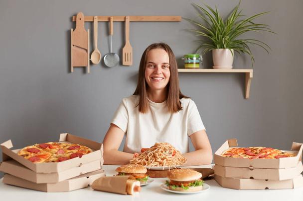 Portrait of joyful cheerful woman with brown hair wearing white T-shirt sitting at table, has tasty dinner with junk food, pizza, hamburgers, sausages in dough, pasta, looking smiling at camera. - Foto, immagini