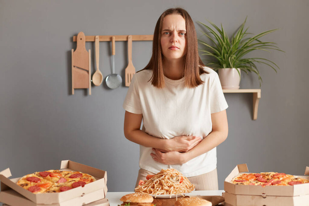 Image of sad ill sick woman with brown hair wearing white T-shirt sitting at table eating junk food and having stomachache, has food poisoning, touching her belly. - Foto, imagen