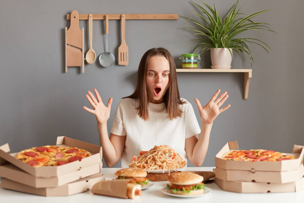 Photo of amazed excited woman with brown hair wearing white T-shirt sitting at table with raised arms and open mouth, being ready to eat pasta, hamburger, pizza and other junk food. - Fotografie, Obrázek