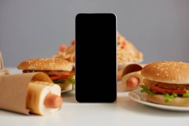 Image of fast food on table, delicious sausage in dough, pasta and meat cutlet burgers, mobile phone with empty display, mockup, advertisement area. - Foto, Imagem