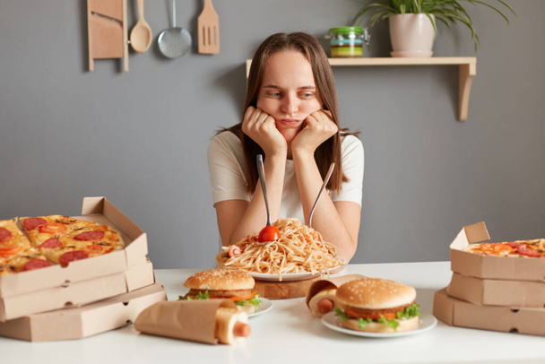Horizontal shot of bored sad upset woman with brown hair wearing white T-shirt sitting at table, keeping diet, looking at fast food and thinking eat or not. - 写真・画像