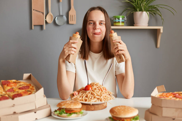 Portrait of hungry woman with brown hair wearing white T-shirt sitting at table eating sausages in dough, showing tongue out, having eating disorder, eating junk food after diet. - Фото, изображение