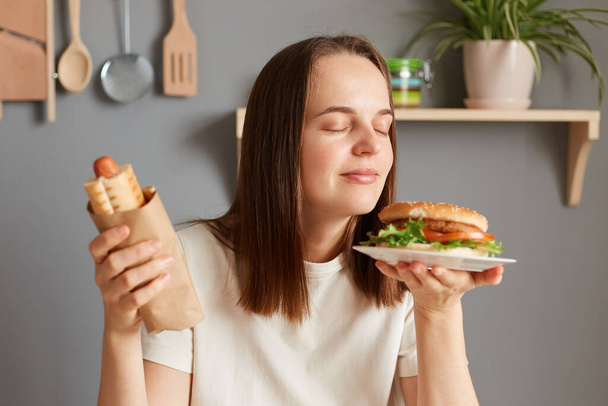 Portrait of joyful satisfied woman with dark hair dressed in white T-shirt holding big burger sandwich and sausage in dough, smelling tasty aromat of junk food, being hungry. - Foto, imagen
