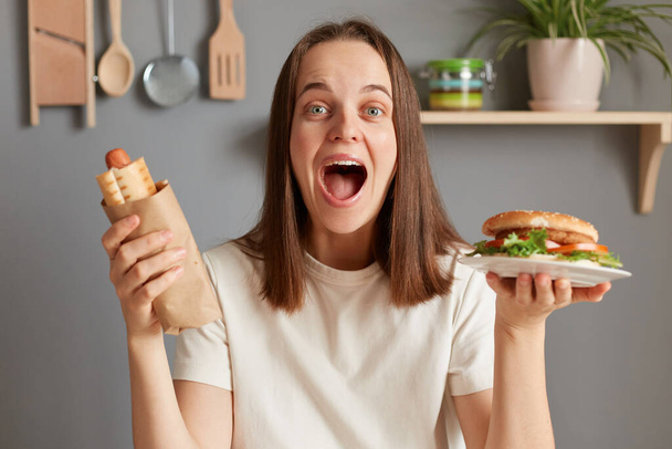 Portrait of extremely happy amazed woman with dark hair dressed in white T-shirt holding big burger sandwich and sausage in dough, rejoicing the end of diet, wants to eat junk food. - Photo, Image