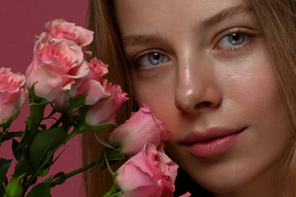 een schoonheid blonde vrouw portret, een roze bloemen in de hand in de buurt van gezicht, roos, roze achtergrond, glimlachen, close-up - Foto, afbeelding