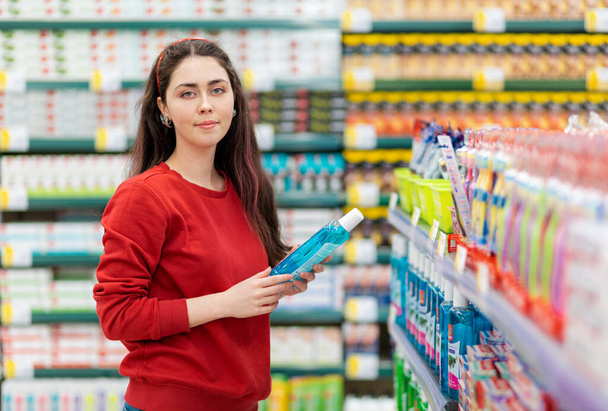 Een jonge blanke vrouw in een rode trui houdt een fles shampoo vast en glimlacht teder. Op de achtergrond, planken met cosmetica. Het concept van het kopen van cosmetica. - Foto, afbeelding