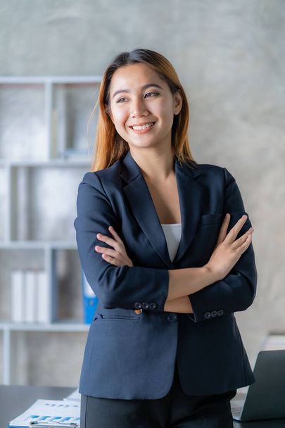 Une jeune comptable asiatique debout les bras croisés devant un bureau avec un ordinateur portable et des documents financiers en arrière-plan au bureau. image verticale - Photo, image