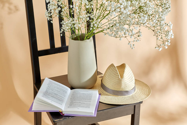 home decor and design concept - close up of gypsophila flowers in vase, book and straw hat on vintage chair over beige background - Φωτογραφία, εικόνα