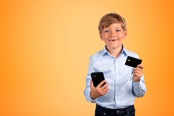 Smiling school boy with smartphone and credit card in hands, portrait on copy space yellow background. Concept of online banking and shopping - Photo, Image