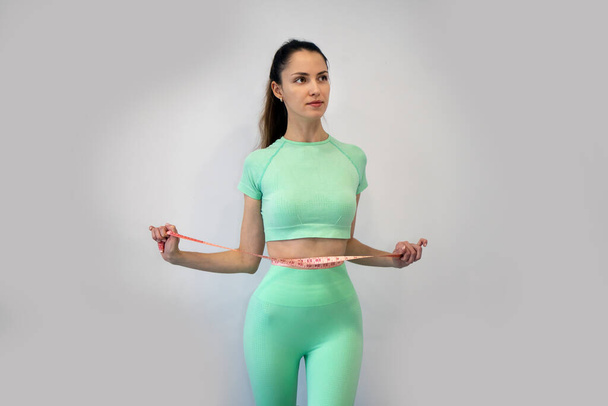Молодая стройная женщина в спортивной одежде измеряет талию рулеткой на сером фоне. Скопируй пространство. Концепция диеты и потери веса - Фото, изображение