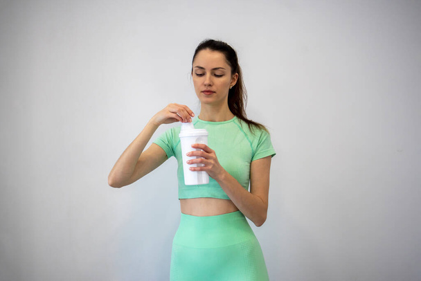 Una donna giovane e magra in abbigliamento sportivo che torce il coperchio di uno shaker nutrizionale sportivo su uno sfondo grigio. Copia spazio - Foto, immagini