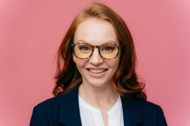 Close up shot of beautiful redhead gorgeous woman in optical glasses, has toothy smile, dressed in elegant clothes, comes on business meeting, happy to sign contract, isolated over rosy background - Photo, Image