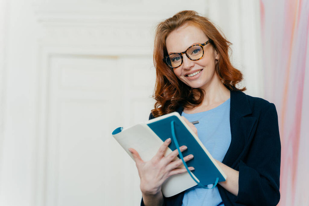 Prosperous satisfied businesswoman in elegant clothes writes in diary, has glad expression, wears spectacles, makes list of plans, writes text message in notebook, stands indoor. Female makes notes - Φωτογραφία, εικόνα