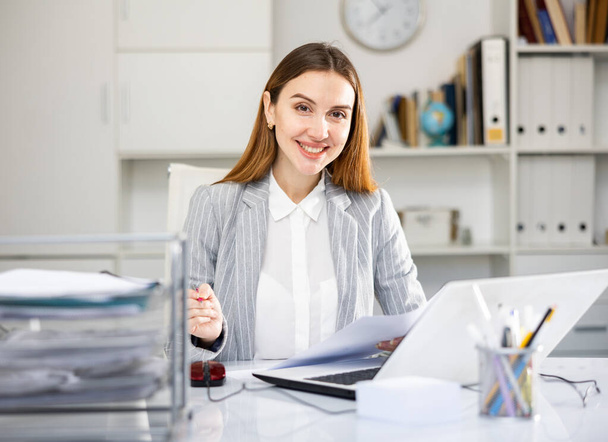 Smiling woman employee working in modern office with documents and laptop, completes a big project - Фото, изображение