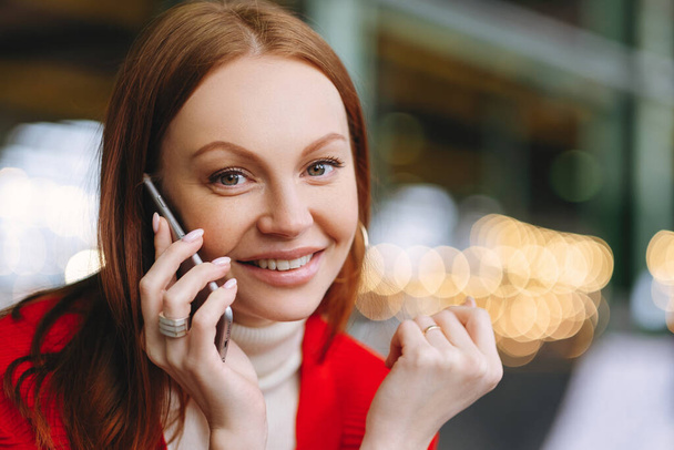 Close up shot of attractive carefree female student talks on mobile phone, enjoys conversation with groupmate, has pleasant smile, poses over blurred background, has spare time after classes - Foto, Imagen