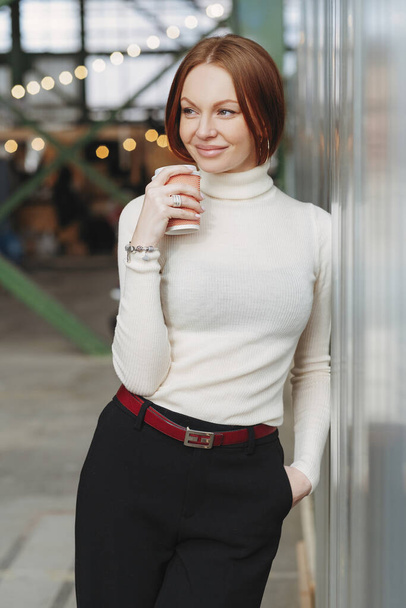 Photo of cheerful young woman wears elegant clothes, dressed in white jumper and black trousers, drinks disposable cup of coffee, dreams about something pleasant, keeps gaze aside. Rest concept - Foto, imagen