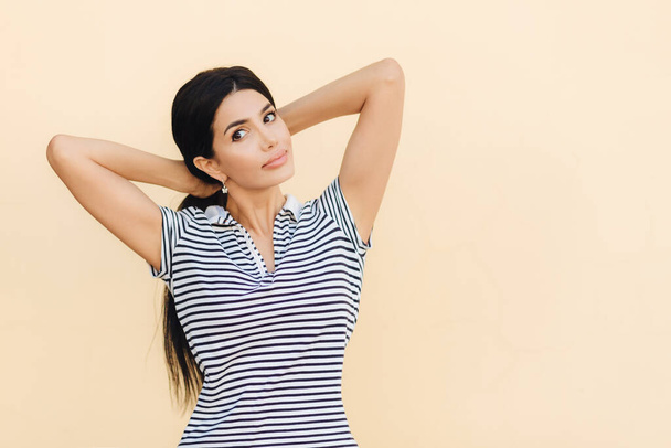 Portrait of pretty female with dark hair, makes pony tail, dressed in casual striped t shirt, looks seriously at camera, poses against studio wall with blank space for your advetisement or promotion - Photo, Image