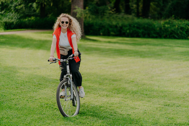 Outdoor image of pretty cheerful young woman rides bike, wears sunglasses, casual wear, poses on green lawn, spends free time in park, bikes in beautiful nature. Activity and recreation concept - Foto, Bild