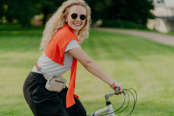 Horizontal shot of happy curly woman rides bike, keeps hands on handlebars, looks active and refreshed, dressed casually, wears shades, poses against green nature background. Spare time concept - Photo, image