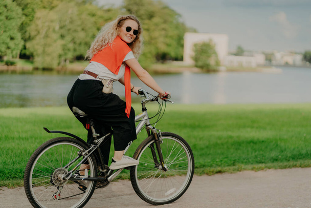 Image of female model rides bicycle, looks aside with cheerful expression, wears sunglasses, breathes fresh air, poses near lake and green trees, covers long distance, finds out something new - Fotó, kép