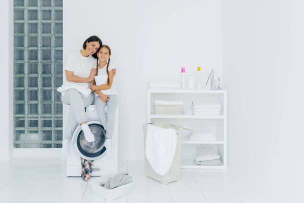 Снимок красивой женщины и его маленькая дочь обнимать и улыбаться приятно, сидеть на стиральной машине, стирать белье в прачечной, иметь дружеские отношения, стирать дома. Концепция домашнего хозяйства
 - Фото, изображение