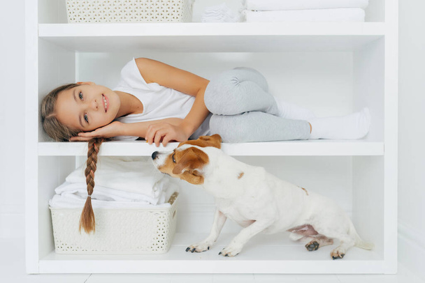 Photo of lovely preschooler lies on white shelf in washing room, has long pigtail, plays with dog, wears casual clothing, smiles gently, spends weekend at home. Children, animals and household concept - Photo, Image