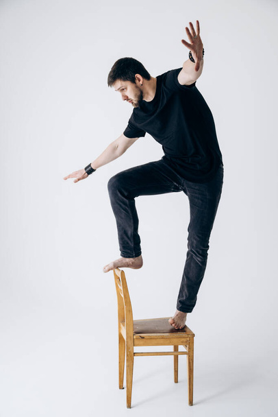 Ο τύπος με το μαύρο μπλουζάκι στέκεται στην καρέκλα και χάνει την ισορροπία του. - Φωτογραφία, εικόνα