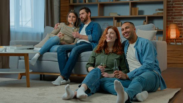 Two ethnic couple diverse men women watch television romantic tv film at home family rest on sofa and floor multiracial friends watching comedy movie comfortable leisure in apartment with bottled beer - Photo, Image