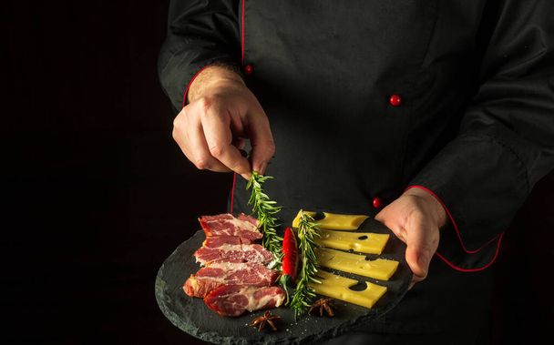 El chef pone romero en un plato con jamón y queso en rodajas. El concepto de servir platos a pedido por un camarero con un lugar para la publicidad sobre un fondo negro - Foto, Imagen