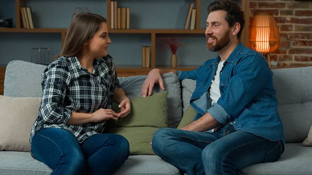 Young couple caucasian hispanic partners husband and wife sit on sofa female woman talk with male friend bearded man talking indoors gossiping friendly dialogue discussing future plans together family - Photo, image