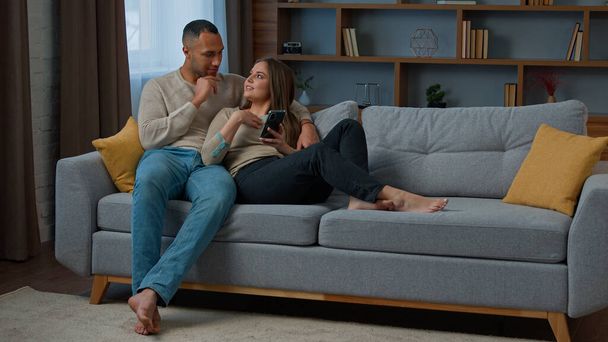 Millennial 30s couple rest on sofa using mobile phone make remote purchase online services choose apartment goods diverse African American man husband and caucasian woman wife browsing home internet - Fotografie, Obrázek