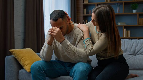 Offended African american man sit on couch upset with bad news health problem Caucasian woman consoling apologize ask forgiveness after quarrel wife touching husband calming support family conflict - Foto, Imagem
