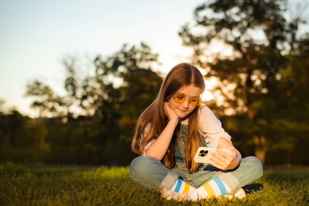 Adorable child girl play and watch video with cell phone sitting in deep grass in park - Foto, afbeelding