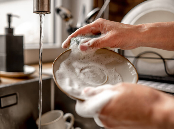 Girl washing plate dish with sponge and foam at kitchen. Woman cleaning utensil with water in sink during daily rouitine - Photo, image