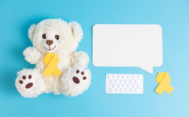 Een speelgoedbeer met witte teddybeer met gele papieren linten, een wit pakje met pillen en een lege callout met ruimte voor tekst ligt op een blauwe achtergrond, vlak van dichtbij. Concept voor Wereld Kinderkankerdag. - Foto, afbeelding